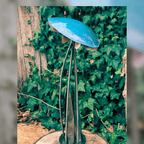 Metallic sculpture of a mushroom with a blue top and metal rods for a base, against a green leafy background