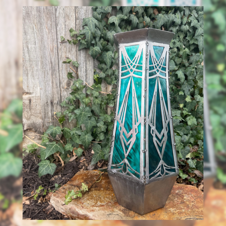 Decorative metal vase with a glass lantern look with geometric design on a stone surface with greenery in the background