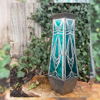 Decorative metal vase with a glass lantern look with geometric design on a stone surface with greenery in the background