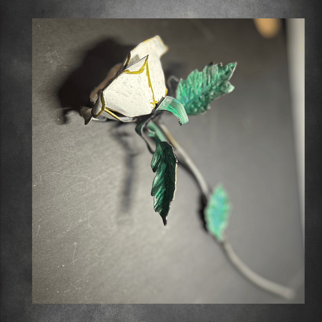 White and gold metal rose side view, at an angle, with green metal leaves on a dark surface